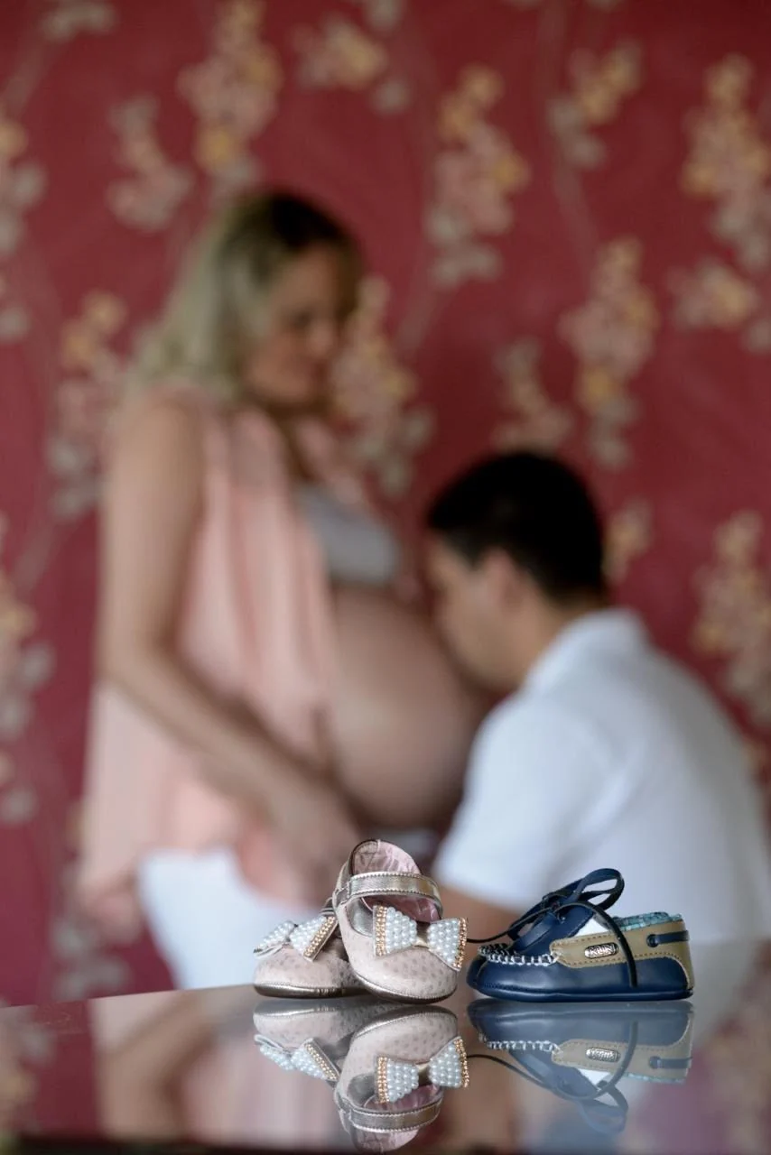 Keila grávida e Gledson ao fundo; em primeiro plano, dois pares de sapatinhos de bebê.